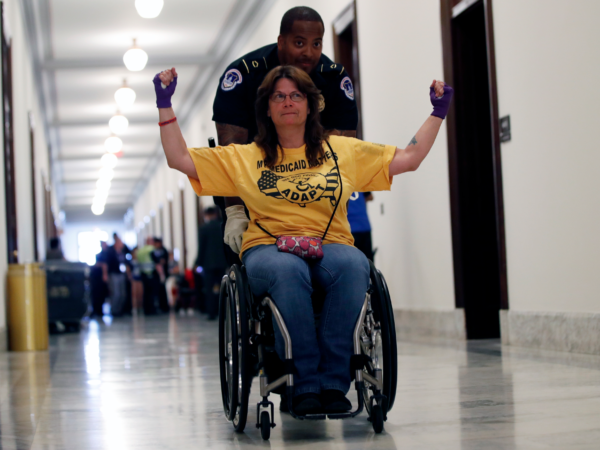 Figure 3: AP Photo/ Jacquelyn Martin. Caption: A protester is removed from a sit-in outside Mitch McConnell’s office on Capitol Hill in June 2017. (AP Photo / Jacquelyn Martin) Image description: A white woman with long brown hair wearing a yellow ADAPT shirt, jeans, and purple fingerless gloves sits in her wheelchair. Her arms are raised to shoulder height and her elbows are bent at right angles (a pose of defiance and victory?). An African-American U.S. Capitol Police officer in his uniform pushes her away from the scene of an ADAPT protest. His latex-gloved right hand is visible under the woman’s right shoulder as he grasps her chair. They are rolling down a white, marble-tiled hallway. There is a group of people, very out of focus, in the background. Both the protestor and the officer have half-smiles on their faces.