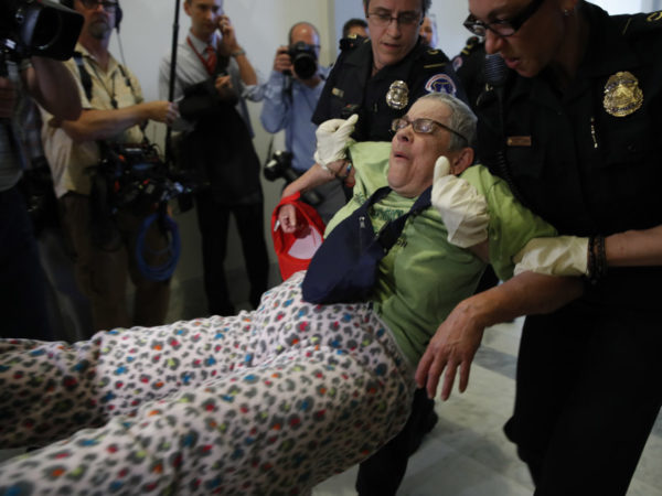 Figure 1: AP Photo / Jacquelyn Martin. Caption: “People are removed from a sit-in outside of Senate Majority Leader Mitch McConnell’s office as they protest proposed cuts to Medicaid, Thursday, June 22, 2017 on Capitol Hill in Washington.” Image Description: A white woman (Spitfire Sabel) is held suspended in the air by two U.S. Capitol Police officers wearing loss-fitting latex gloves. In the background are many journalists and other cameras capturing the arrest. They are out of focus while Sabel and the two white, female officers arresting her are in sharp focus.