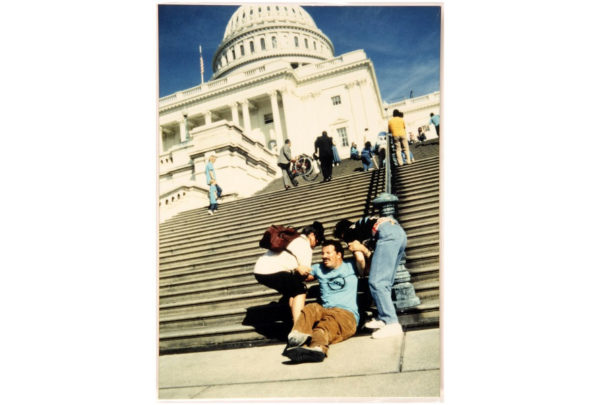 Figure 2: Tom Olin. Caption: “A man (George Roberts) is seated at the bottom of the flight of stairs up to the Capitol building. His feet are cross and legs tense from CP. A couple of standing people are getting ready to assist him by lifting him up the steps. Higher on the flights a woman and a child in light blue ADAPT shirts are heading up the steps. Still higher, a couple of men in suits are lifting a manual wheelchair (without a person in it) up the stairs. Still higher someone on a crutch is climbing the steps and someone else is crawling up by the railing in the middle of the stairs as a camera-man films. Still higher a few others are reaching the top of the flight of stairs. The white Capitol dome and building are set against the blue sky.”