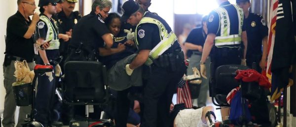 Figure 4: Mark Wilson / Getty Images. Caption: “ADAPT member Anita being carried away at the protest in Senator McConnell’s office, while other members are lying on the ground in the background.” Image description: This wide-angle photograph shows the arrest of Anita, an ADAPT member, at the protest outside Senator McConnell’s office. Anita, an African-American woman, is held above her motorized wheelchair by three U.S. Capitol Police officers. All three officers—a white woman, a white man, and an African American man—wear latex gloves. On the far left side of the photograph there is a line of other officers, one of whom is wearing gloves, and a white man with an ear-piece talking into a small microphone attached to a phone. At the center-right of the photograph, a protestor lies on the floor. Only their right leg is visible and they have a black brace on that leg. A U.S. Capitol Police officer stands over the protestor with his back to the camera, his latex-gloved hands at his sides. The officer’s head is cropped out of the photograph, but he appears to be looking slightly away from the protestor, toward another officer walking toward him. An empty wheelchair sits behind the officer. There is a small U.S. flag at the center of the photograph, right above the protestor. Another U.S. flag and an MIA/POW flag appear on poles on the far right of the photograph. The red and white stripes of what might be another U.S. flag peak out of the bag on the back of the empty wheelchair.