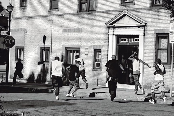 Rochester, New York, USA: Youths run away as a police car approaches a corner known as a drug-dealing spot. The group of deprived neighborhoods known as the ‘Crescent’, around the northern edge of downtown Rochester, New York State, USA, is renowned for its high crime and murder rates. Reasons given for these include a depressed local economy, and the large number of empty houses, prone to becoming locations for drug dealing. Magnum Photos for Postcards from America / Die Zeit Magazine