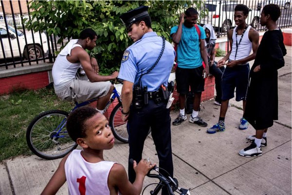 First year officer Jonathan Dedos (a "foot beat") questions a group of young men, after a shooting suspect was described as an African-American wearing white and on a bike, a description which could apply to many young men in the neighborhood. This group, at a local park, was vouched for by a man who works with local youth, and the police moved on. July 28, 2015. Philadelphia, Pa.