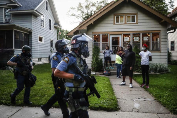 people watching as police in riot gear walk down a residential street, Thursday, May 28, 2020, in St. Paul, Minnesota.